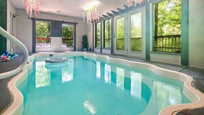 An indoor swimming pool with clear blue water, surrounded by large windows offering views of greenery outside.