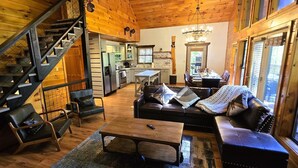 A cozy cabin interior with a wooden staircase, a dining area, a living room with a sofa and coffee table, and large windows.