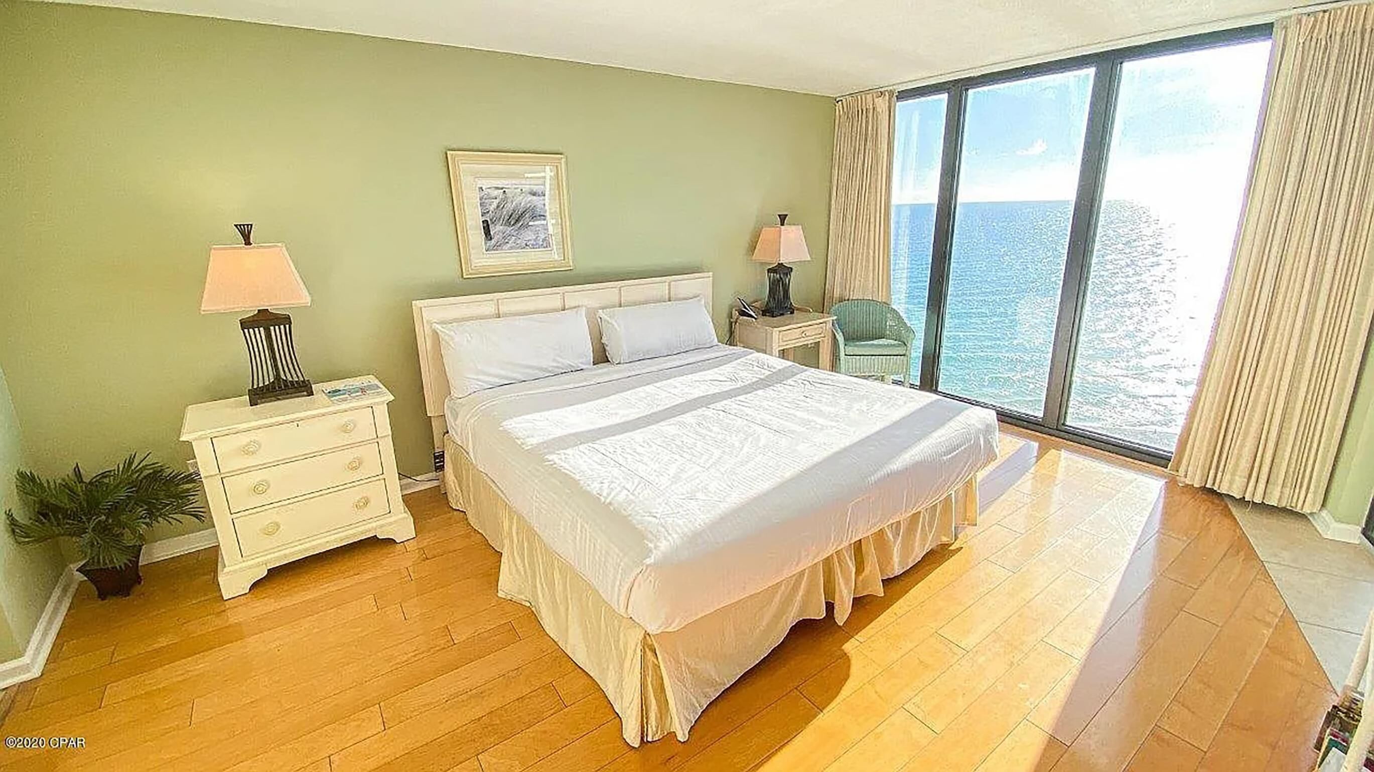Un dormitorio con una cama grande, una mesita de noche, una lámpara, una silla y vistas al mar a través de un ventanal amplio.