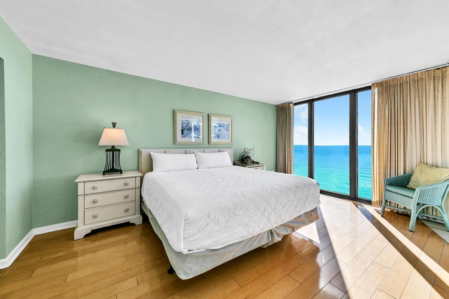 Un dormitorio con una cama grande, una mesita de noche con lámpara, una silla junto a la ventana y vistas al océano.