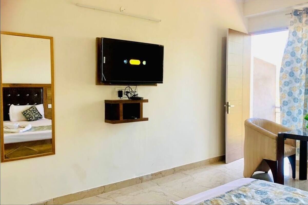 Une chambre d’hôtel avec un lit, un téléviseur fixé au mur, une petite table avec une chaise et un balcon agrémenté de rideaux à motifs.