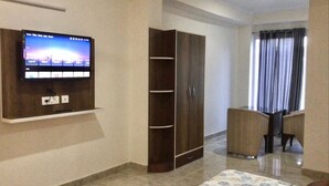 Una habitación con televisión de pantalla plana en la pared, un armario, una zona de comedor con dos sillas y una ventana con cortinas.