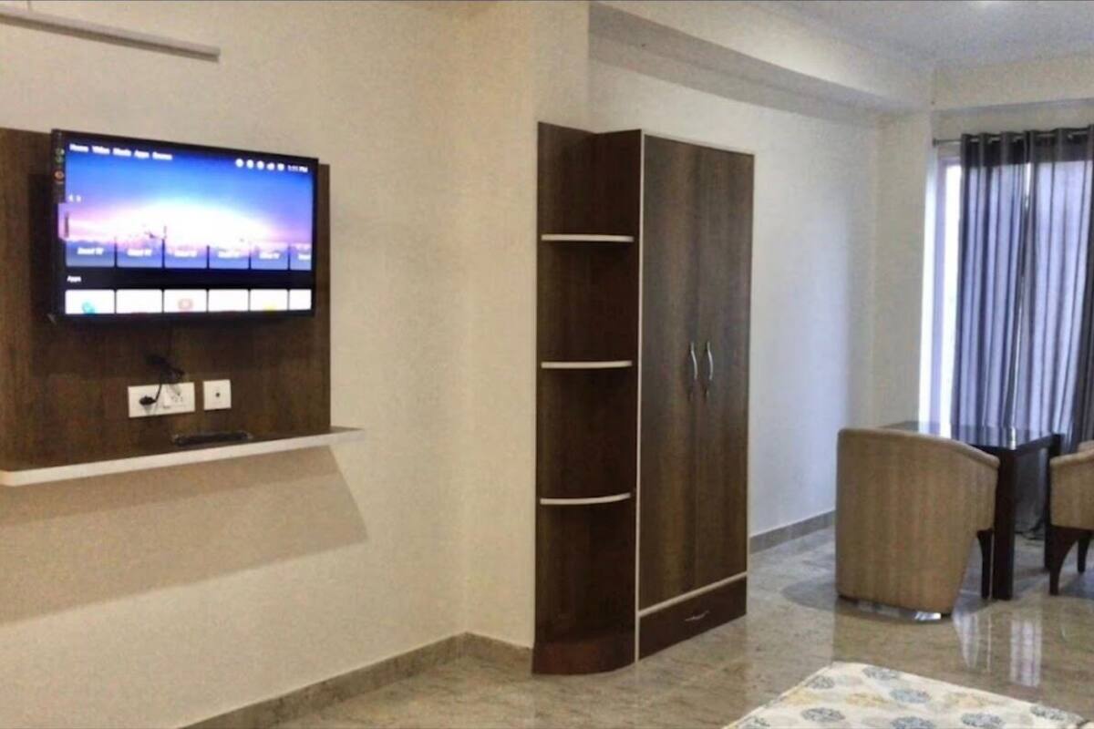 Une chambre dotée d’un téléviseur à écran plat fixé au mur, d’une armoire, d’un coin repas avec deux chaises et d’une fenêtre avec des rideaux.