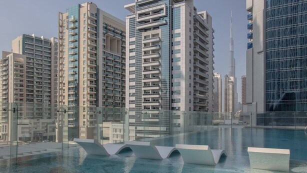 A modern residential building with a glass facade, a rooftop pool with lounge chairs, and a clear view of the surrounding high-rise buildings.