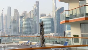 Un área de piscina en la azotea con sillas de descanso y una estatua, con vistas a un horizonte urbano con edificios modernos.