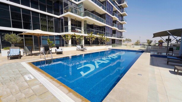Ein Swimmingpool mit dem Schriftzug "DAMAS" auf der Wasseroberfläche, umgeben von Liegestühlen und Sonnenschirmen, mit einem modernen Gebäude im Hintergrund.
