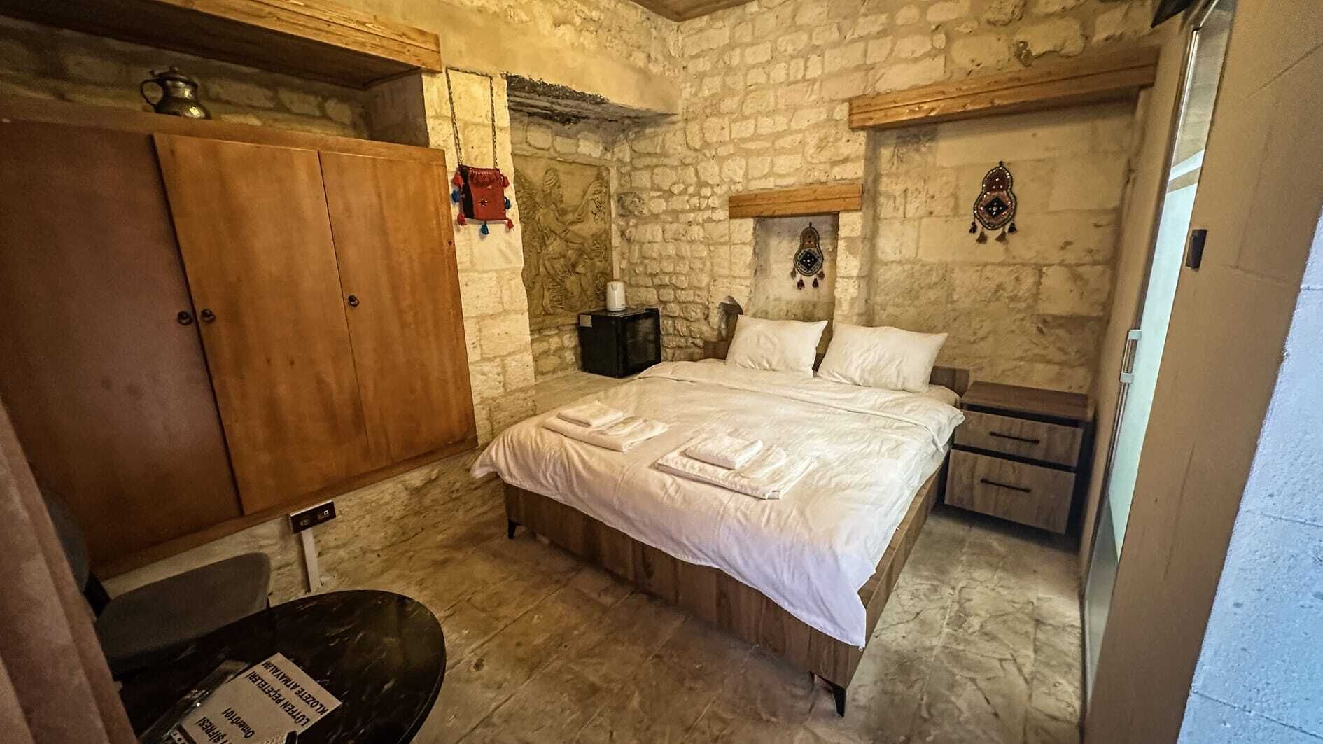 A bedroom with a stone wall, wooden bed frame, and a nightstand with a lamp.