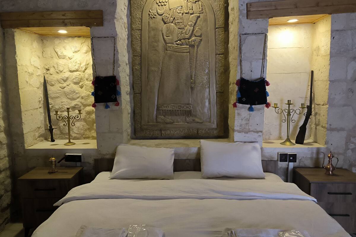 A bedroom with a large bed, two bedside tables, a stone wall, and a carved wooden panel above the bed.