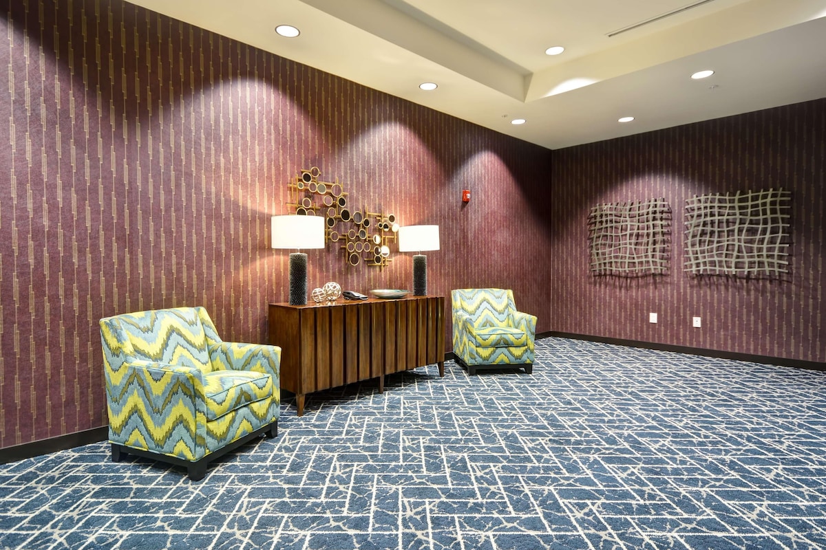 Una habitación con papel tapiz estampado, una consola de madera con lámparas, dos sillones estampados y una alfombra con diseño.
