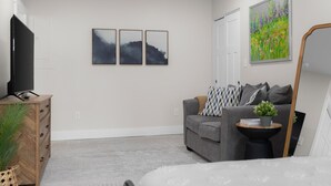 Una sala de estar moderna con un sofá gris, un televisor de pantalla plana sobre un mueble de madera, una mesita auxiliar con una planta y un espejo grande.