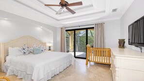 A bedroom with a bed, a nightstand, a desk, a chair, a ceiling fan, and a large window with curtains.