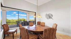 Un espace repas avec une grande table ronde entourée de chaises en osier, une vue sur l’extérieur grâce à de grandes fenêtres et un élément décoratif mural.