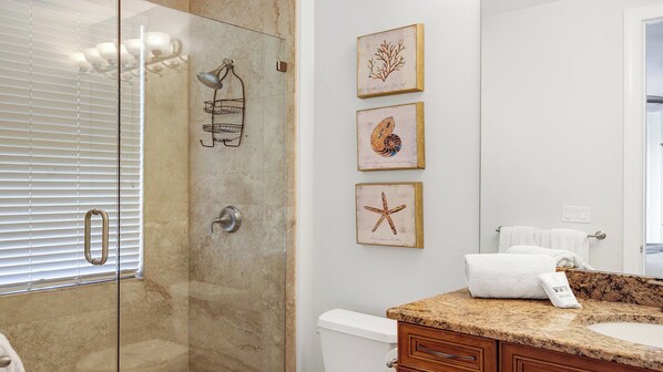 A bathroom with a glass-enclosed shower, a granite countertop, a white toilet, and three framed sea-themed pictures on the wall.