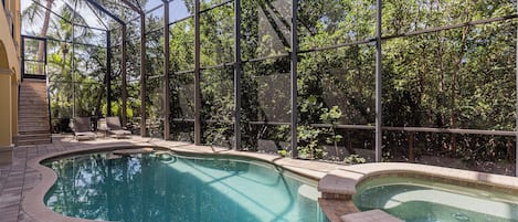 A swimming pool with a glass enclosure, surrounded by trees and a patio area with a chair and a small staircase.
