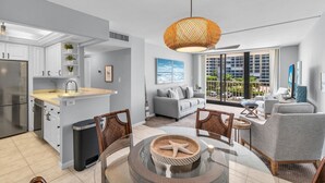 Un appartement moderne comprenant une cuisine, un coin repas et un salon. Il dispose d’une grande fenêtre offrant une vue sur la ville, d’une table à manger avec des chaises et d’un canapé.
