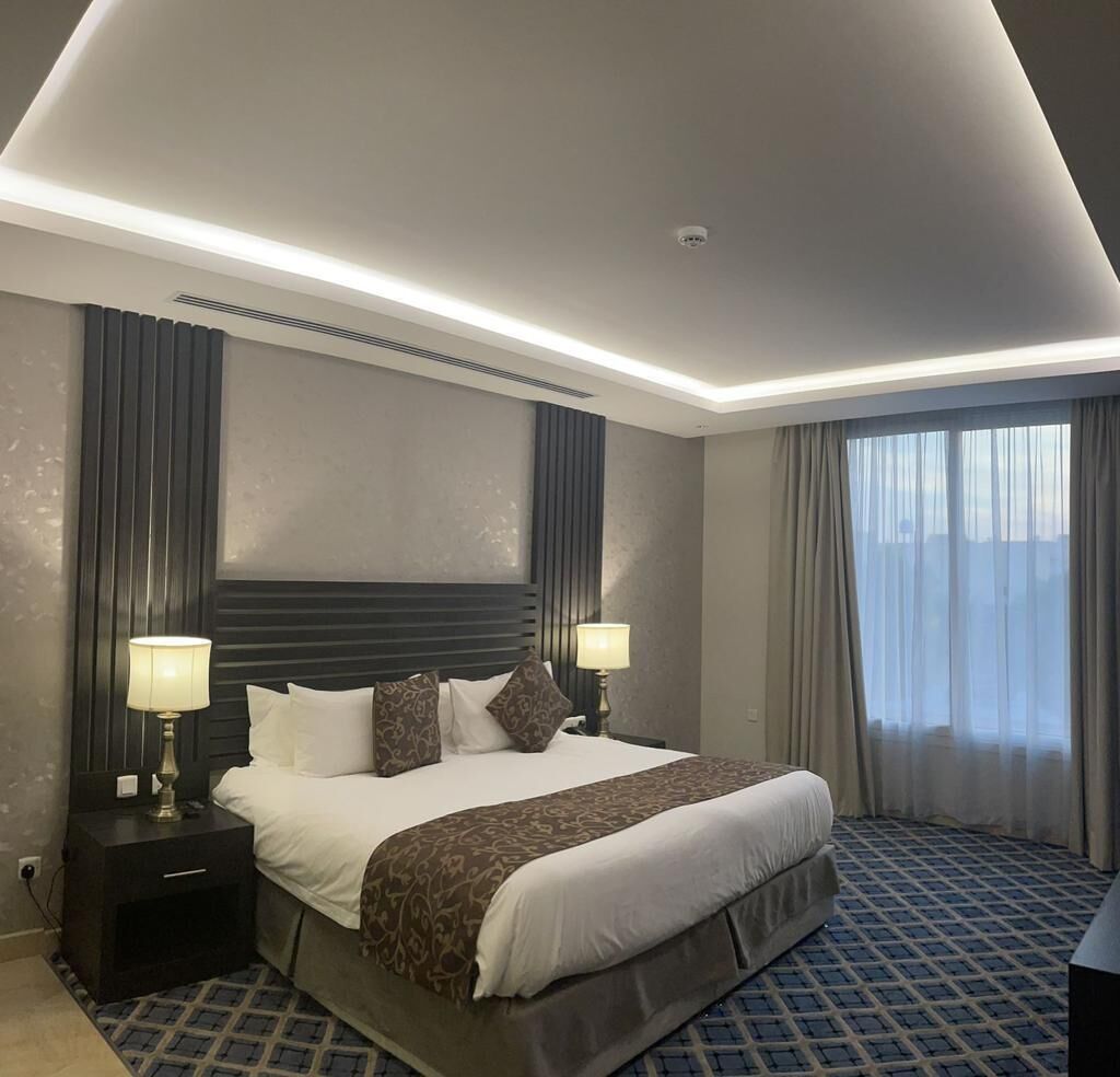 Una habitación de hotel moderna con una cama grande, dos mesitas de noche con lámparas, un cabecero y un amplio ventanal con cortinas.