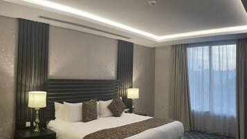 Una habitación de hotel moderna con una cama grande, dos mesitas de noche con lámparas, un cabecero y un amplio ventanal con cortinas.