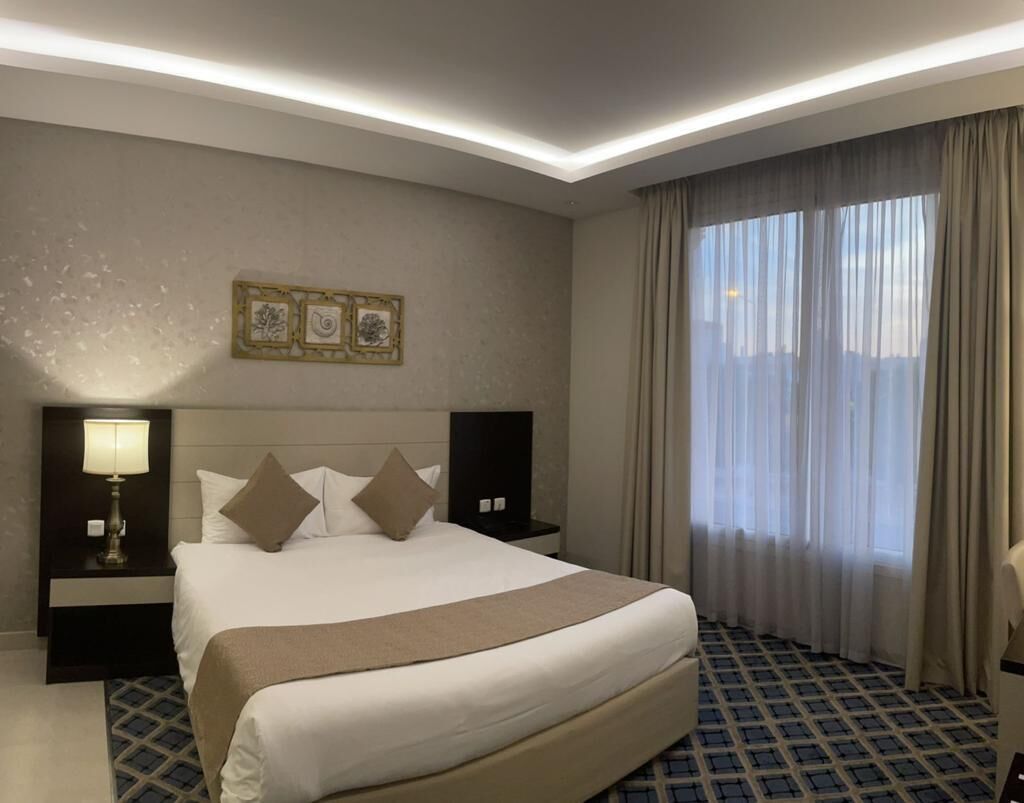 A hotel room with a large bed, a nightstand with a lamp, a wall-mounted TV, and a window with curtains.