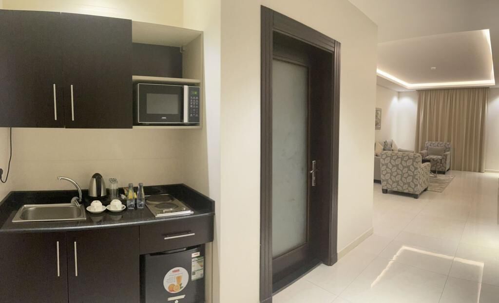 Cocina compacta con microondas, pileta y heladera pequeña. Una puerta oscura da a una sala de estar con sofá y alfombra estampada.
