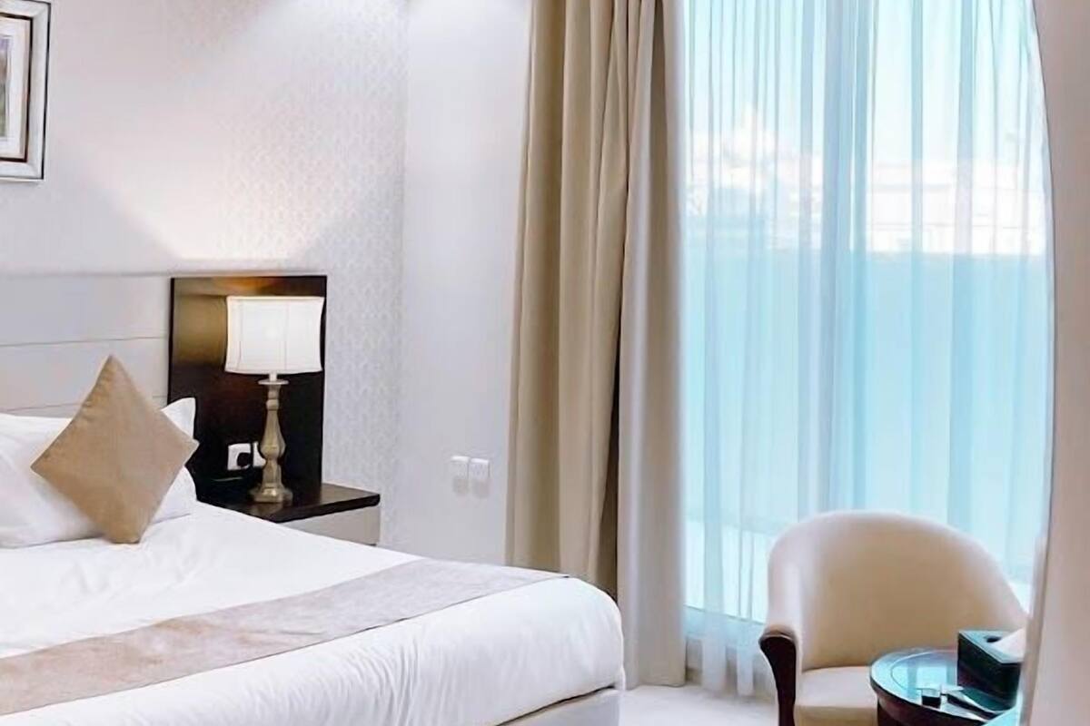 A modern hotel room with a large bed, a desk, a chair, and a view of the sea through sheer curtains.