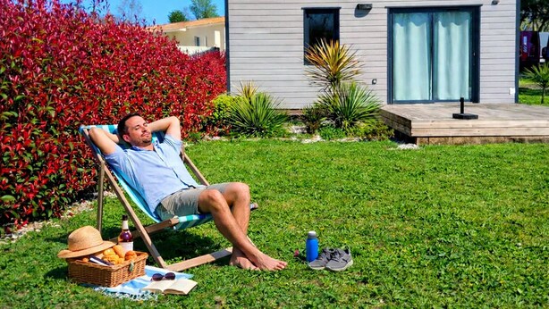 Una persona descansando en una silla de jardín con una cesta de comida y bebidas cerca, frente a una casa moderna con un pequeño jardín.