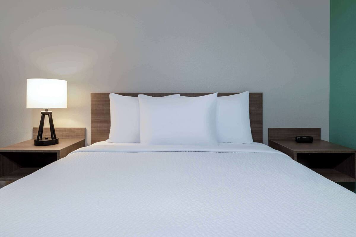 Uma cama bem arrumada com lençóis brancos, cabeceira de madeira e uma luminária de cabeceira em um criado-mudo.