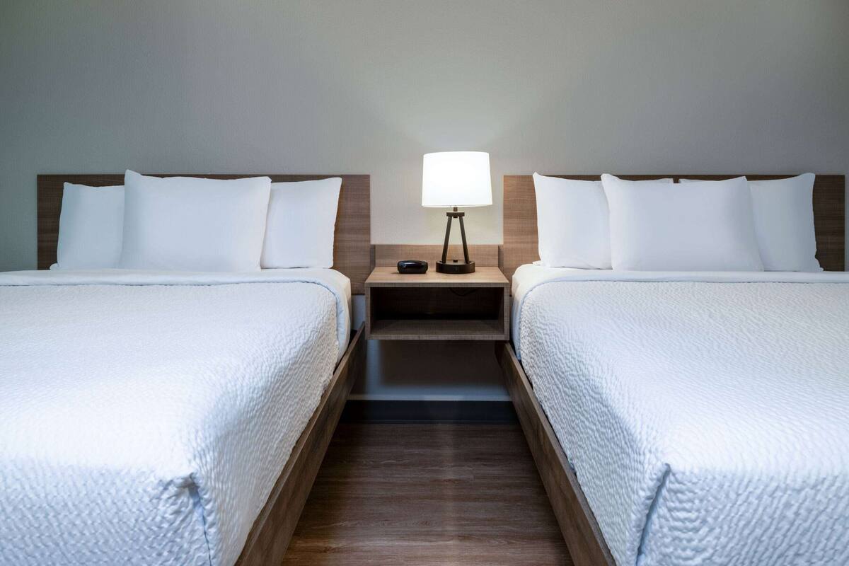 Quarto de hotel com duas camas, uma mesa de cabeceira com abajur e uma cabeceira de madeira.