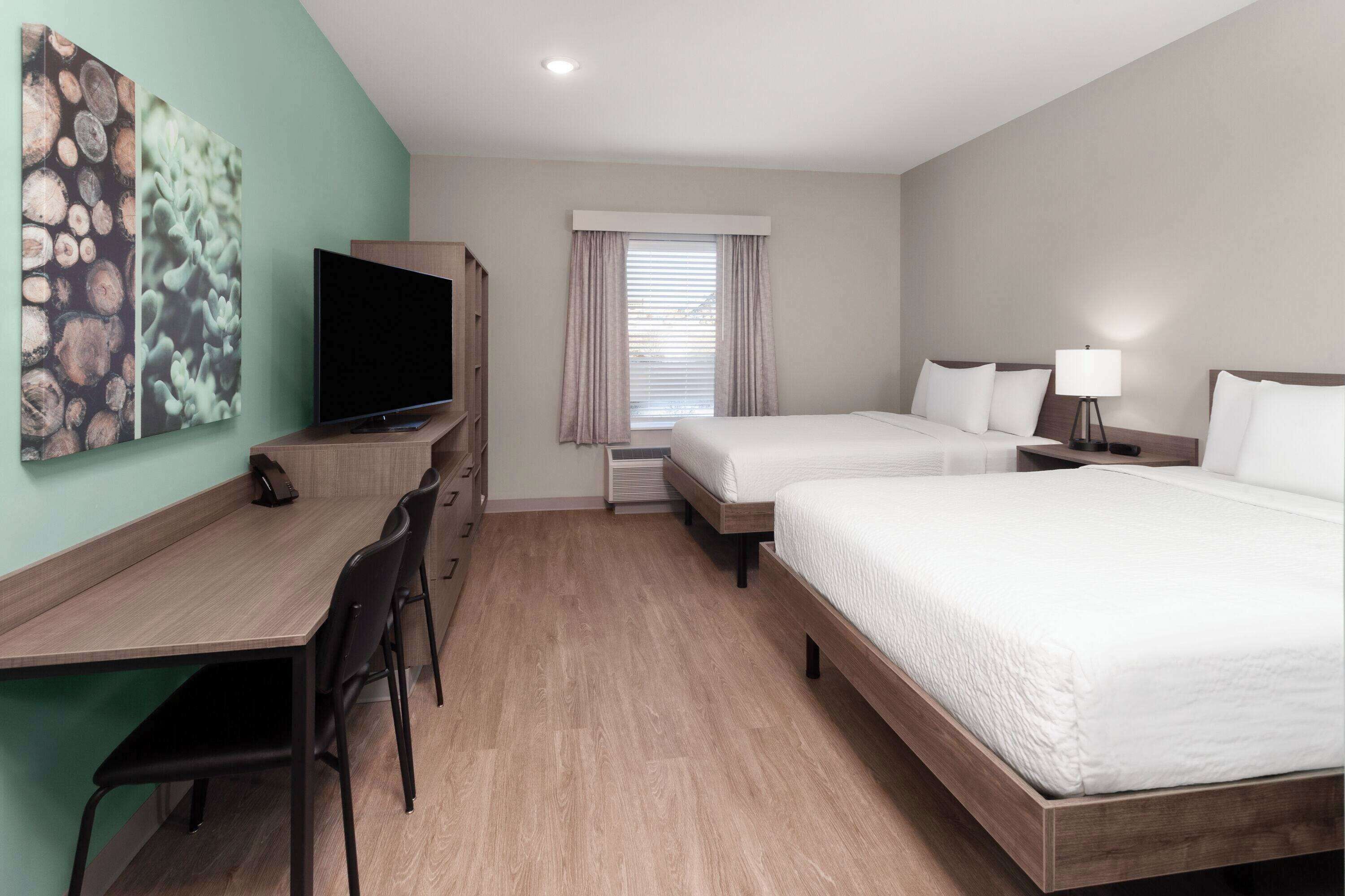 A hotel room with a wooden desk, two beds, a TV, and a window with curtains.