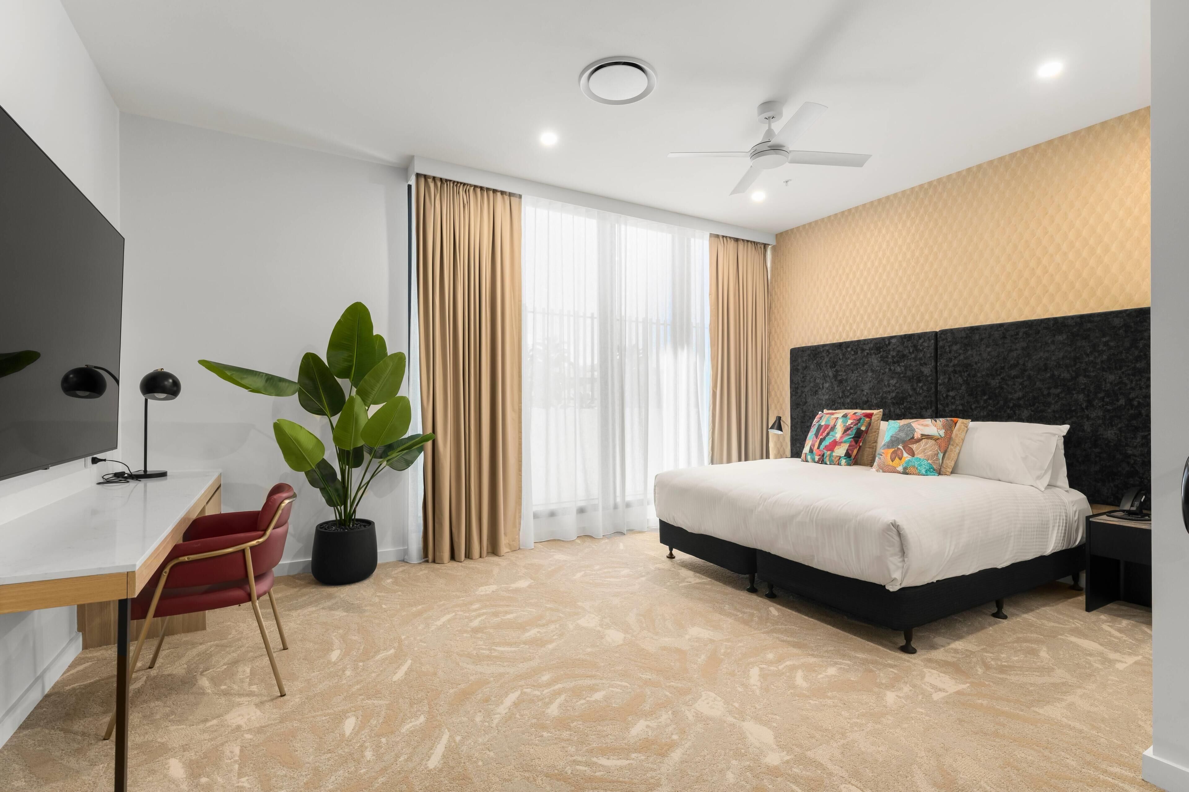Quarto de hotel moderno com cama, escrivaninha, cadeira, planta em vaso e uma grande janela com cortinas.