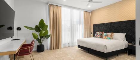 Quarto de hotel moderno com cama, escrivaninha, cadeira, planta em vaso e uma grande janela com cortinas.