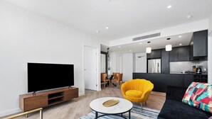 A modern living room with a flat-screen TV, a sofa, a yellow armchair, and a kitchen area in the background.