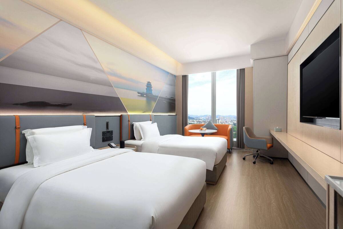 Een hotelkamer met twee bedden, een bureau, een stoel, een televisie en een groot raam met uitzicht op de stad.