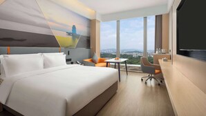 A hotel room with a large bed, a desk, a chair, and a TV. There is a city view through the windows.