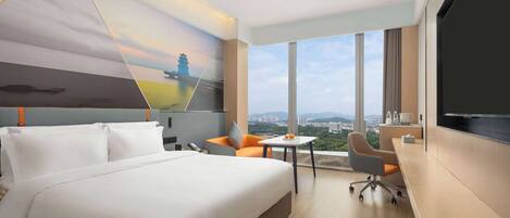 A hotel room with a large bed, a desk, a chair, and a TV. There is a city view through the windows.