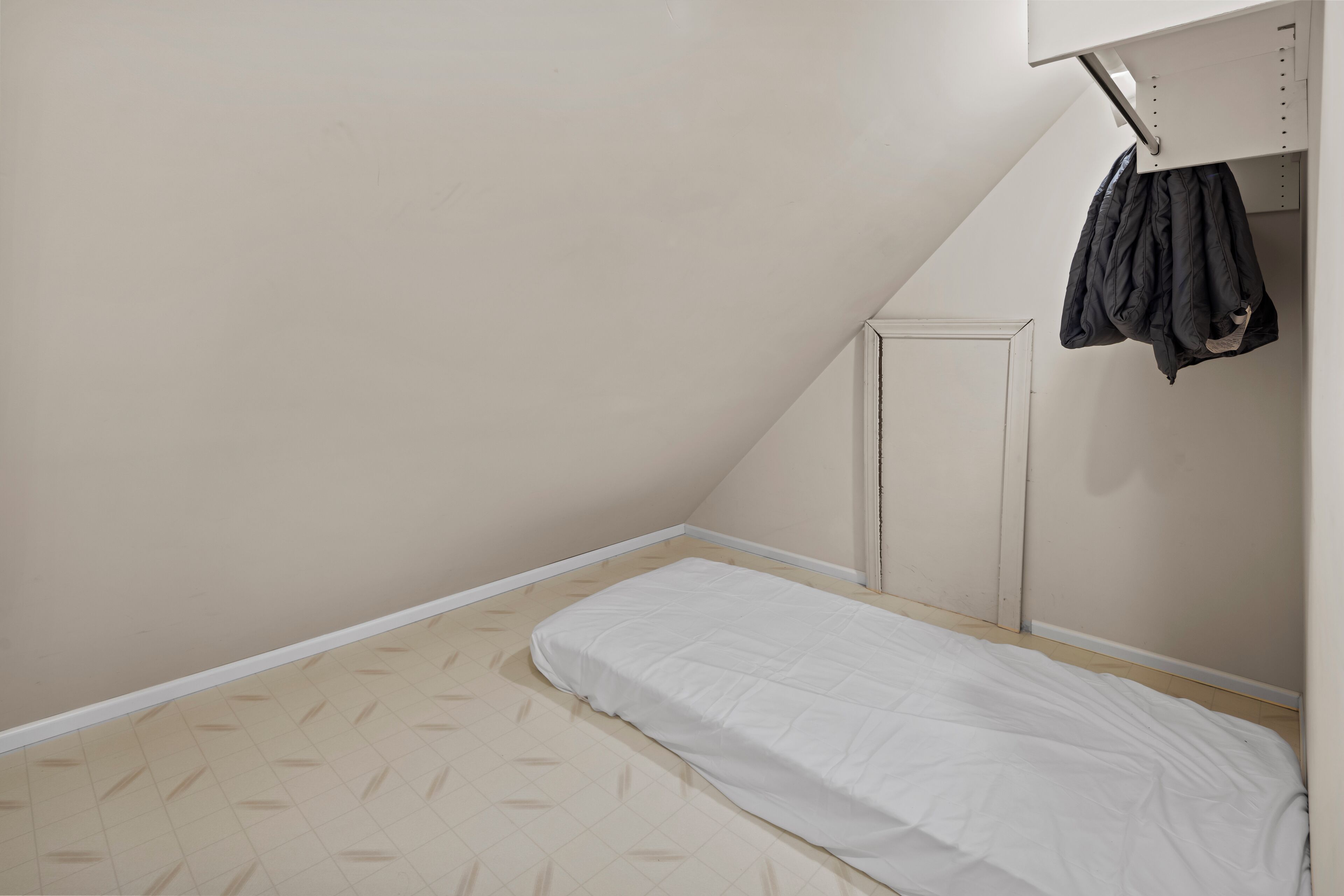 A small, single-bed room with a white mattress, a slanted ceiling, and a coat hanging on a rack.