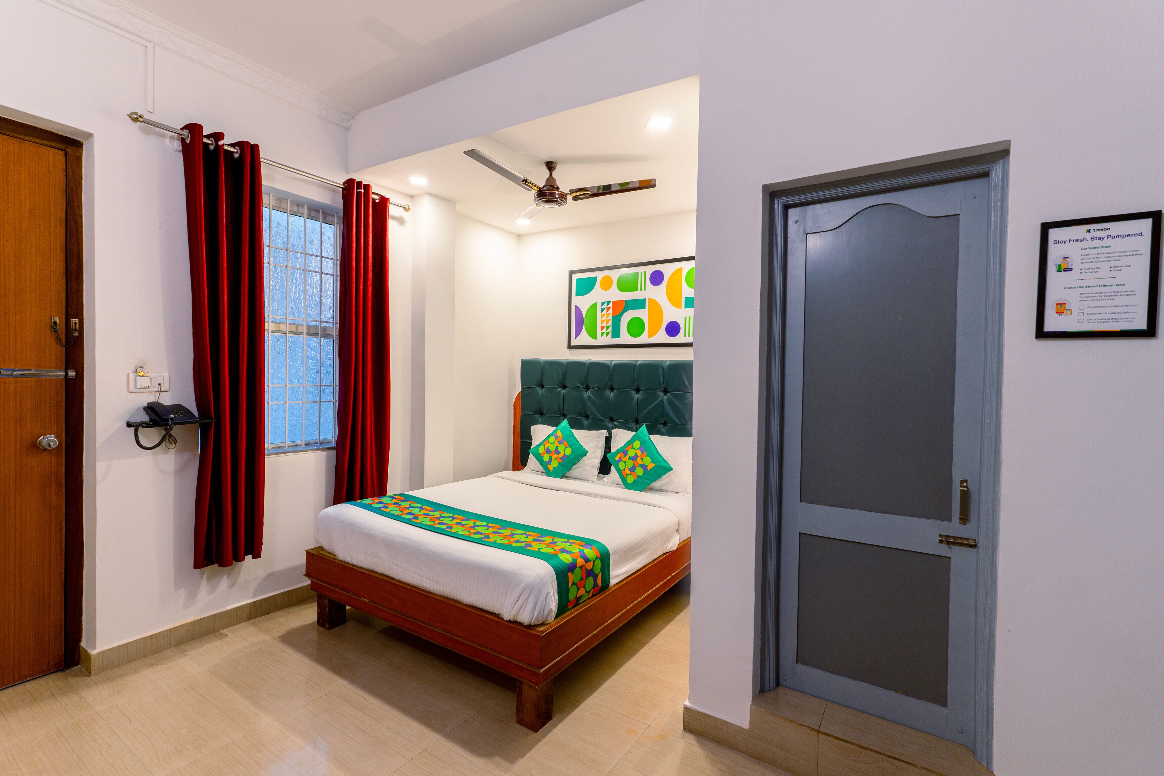 Une chambre d’hôtel avec un lit, un ventilateur de plafond, une fenêtre avec des rideaux, une porte et un panneau fixé au mur.