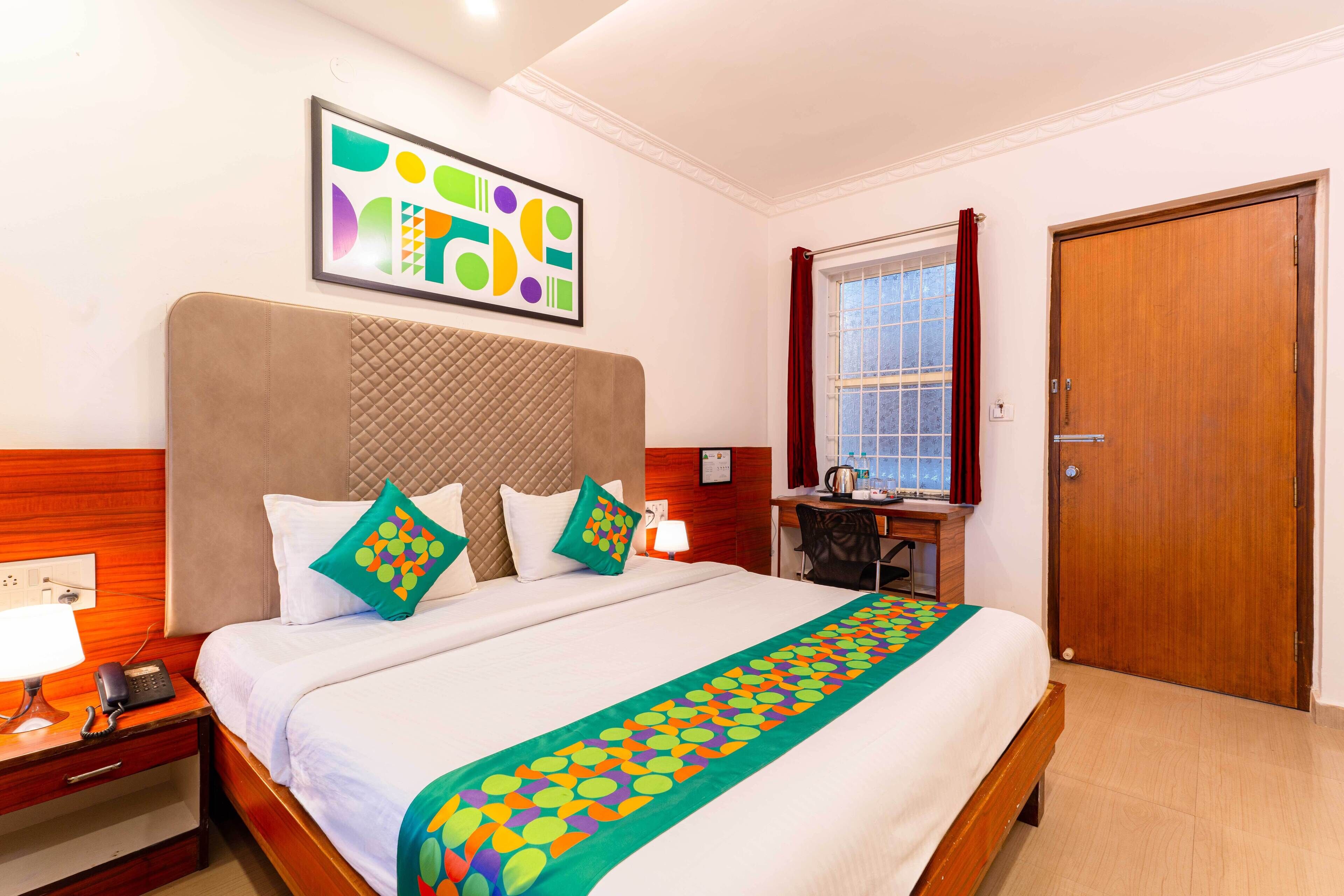 A hotel room with a large bed, a desk, a chair, a window with curtains, and a door.