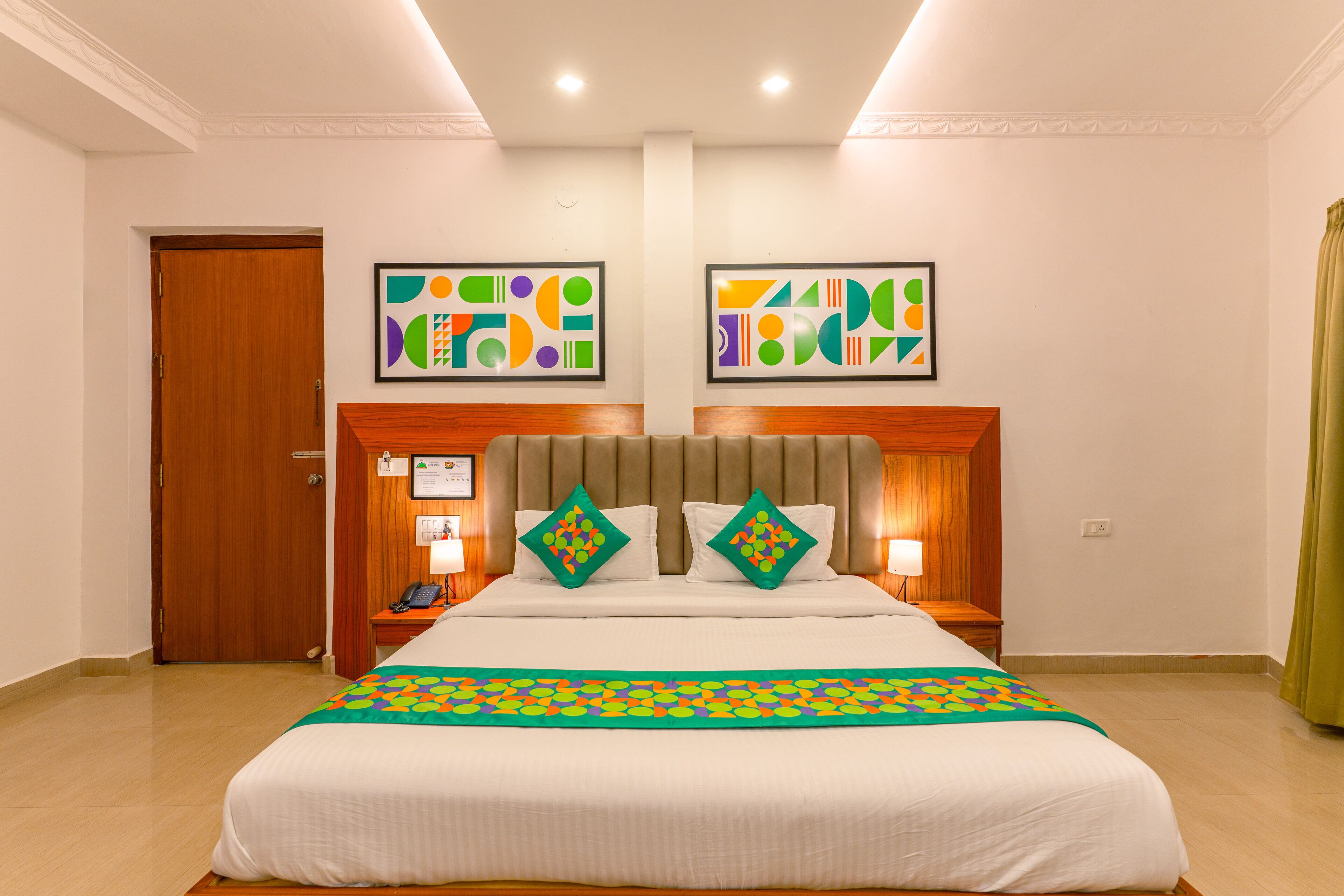 A hotel room with a large bed, two bedside tables, a wooden door, and two framed abstract paintings on the wall.