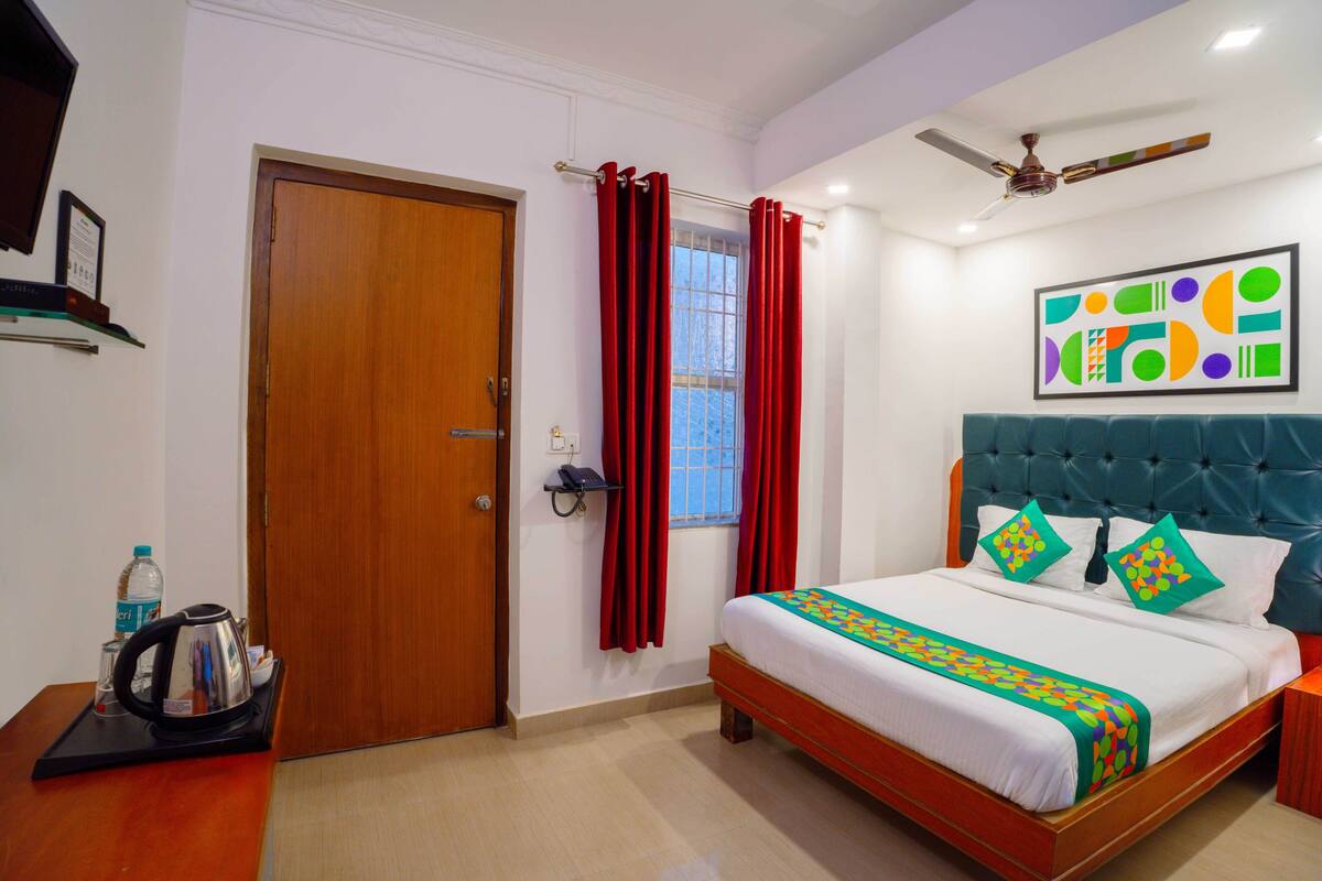 A hotel room with a wooden door, a bed with a blue headboard, a ceiling fan, and a window with red curtains.