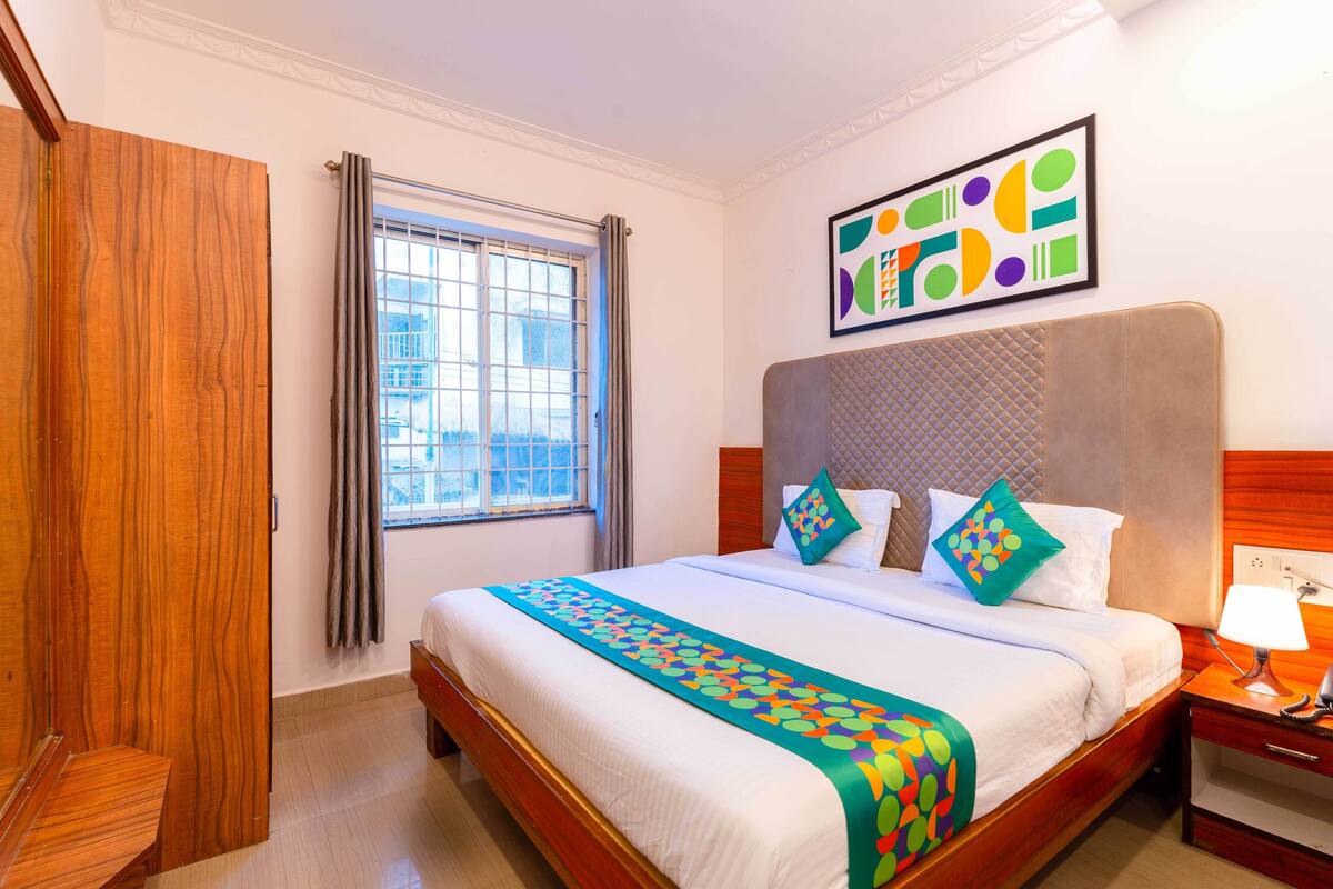 A hotel room with a large bed, a wooden wardrobe, a window with curtains, and a colorful abstract painting on the wall.