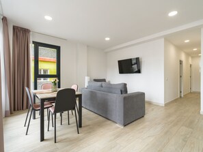 A modern living room with a grey sofa, a dining table with chairs, and a flat-screen TV mounted on the wall.