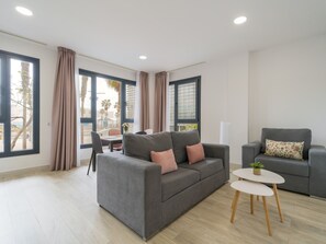 Un soggiorno moderno con un divano grigio, una poltrona grigia più piccola e un tavolino bianco rotondo con una pianta. Ampie finestre con tende permettono alla luce naturale di illuminare l'ambiente e offrono vista sull'esterno.