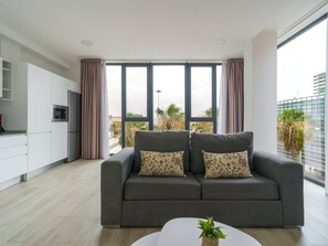 Uma sala de estar moderna com um sofá cinza, uma mesinha branca e uma janela grande com cortinas.