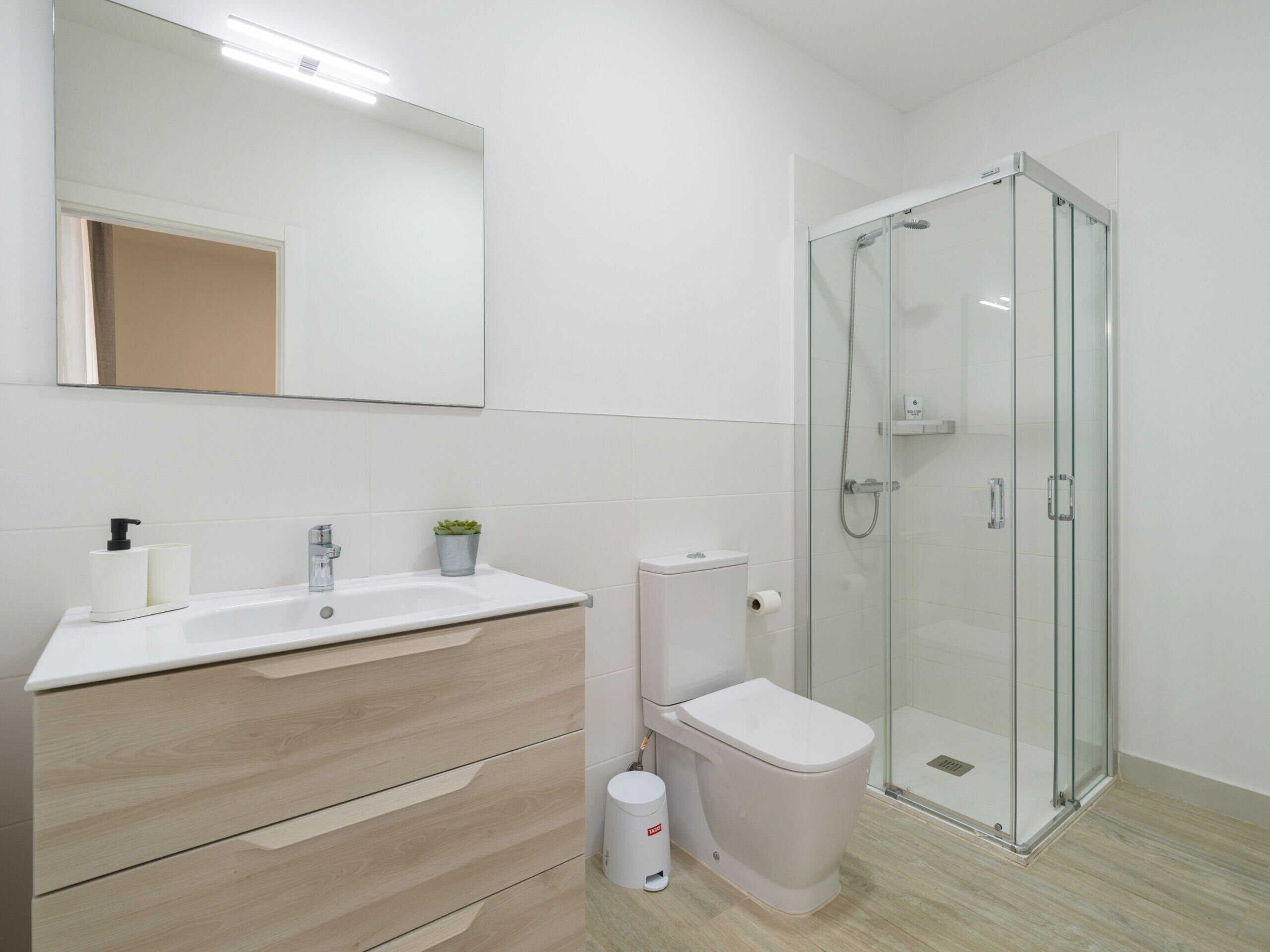 Un baño moderno con un lavabo blanco, inodoro, cabina de ducha con mampara de vidrio y espejo sobre el lavabo.