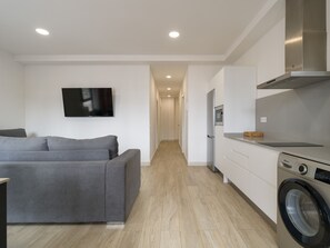 Un appartamento moderno con un divano grigio, una TV a schermo piatto, una lavatrice e una cucina con mobili bianchi.