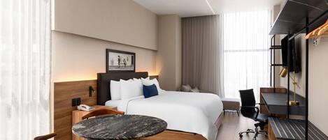 A modern hotel room with a bed, bedside table, chair, desk, and a small round table.