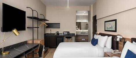 A hotel room with two beds, a desk with a computer, a TV, a small kitchenette, and a bathroom.