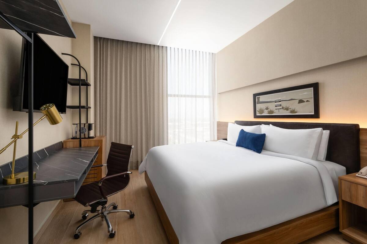 A hotel room with a bed, a desk with a chair, a TV mounted on the wall, and a window with curtains.