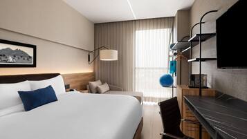 A modern hotel room with a large bed, a desk, a chair, a TV, and a window with curtains.