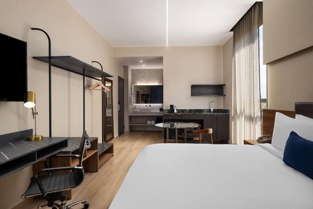 A hotel room with a bed, desk, chair, and a small kitchenette.